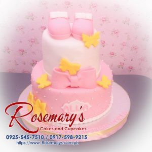 Delicious 2 layer pink and yellow themed cake adorned with baby shoes and a ribbon, perfect for a christening. Rosemary's Cakes, 0925-545-7510.