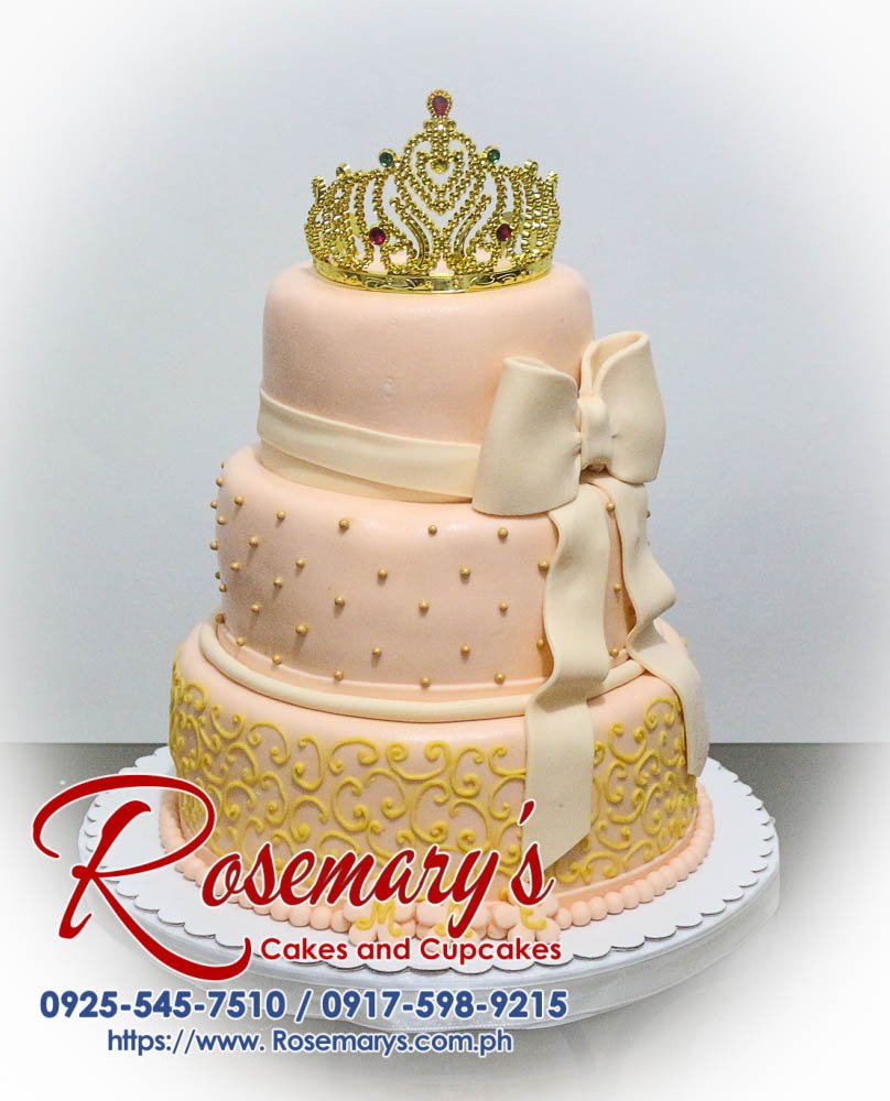 Elegant 3-tier beige cake adorned with a gold crown and delicate ribbon detail. By Rosemary's Cakes. Call 0917-598-9215.